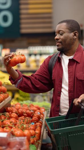 Man in supermarkt bekijkt tros tomaten in groenteschap met boodschappenmandje in zijn andere hand