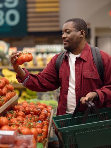 Man in supermarkt bekijkt tros tomaten in groenteschap met boodschappenmandje in zijn andere hand