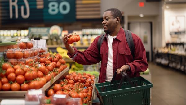 Man in supermarkt bekijkt tros tomaten in groenteschap met boodschappenmandje in zijn andere hand