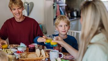 Twee ouders en een kind zitten samen aan tafel te ontbijten, de moeder en het kind pakken allebei sinaasappelsap en kijken elkaar aan
