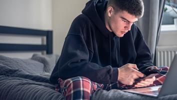 Een jongere zit alleen op zijn bed naar zijn telefoon en laptop te kijken