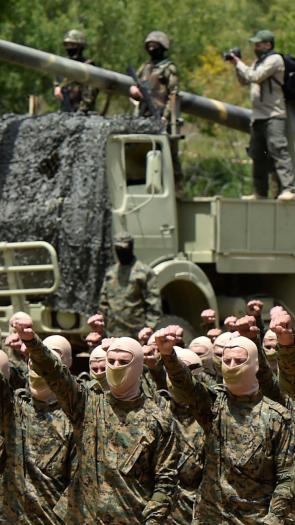 Hezbollah-strijders voeren een militaire oefening uit. Ze staan op een rij in militaire kledin gmet bivakmutsen met de vuisten omhoog. Op de achtergrond wagens en wapens.