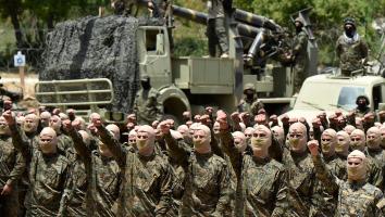 Hezbollah-strijders voeren een militaire oefening uit. Ze staan op een rij in militaire kledin gmet bivakmutsen met de vuisten omhoog. Op de achtergrond wagens en wapens.