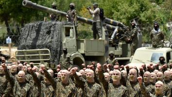 Hezbollah-strijders voeren een militaire oefening uit. Ze staan op een rij in militaire kledin gmet bivakmutsen met de vuisten omhoog. Op de achtergrond wagens en wapens.