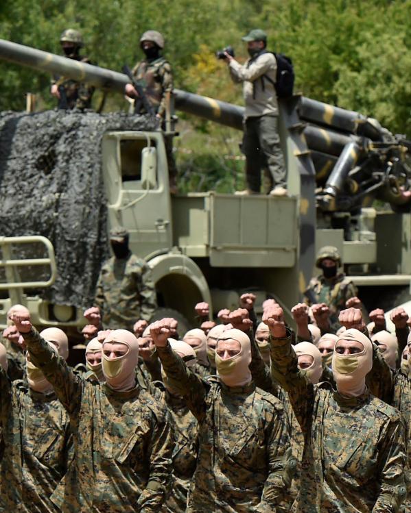 Hezbollah-strijders voeren een militaire oefening uit. Ze staan op een rij in militaire kledin gmet bivakmutsen met de vuisten omhoog. Op de achtergrond wagens en wapens.