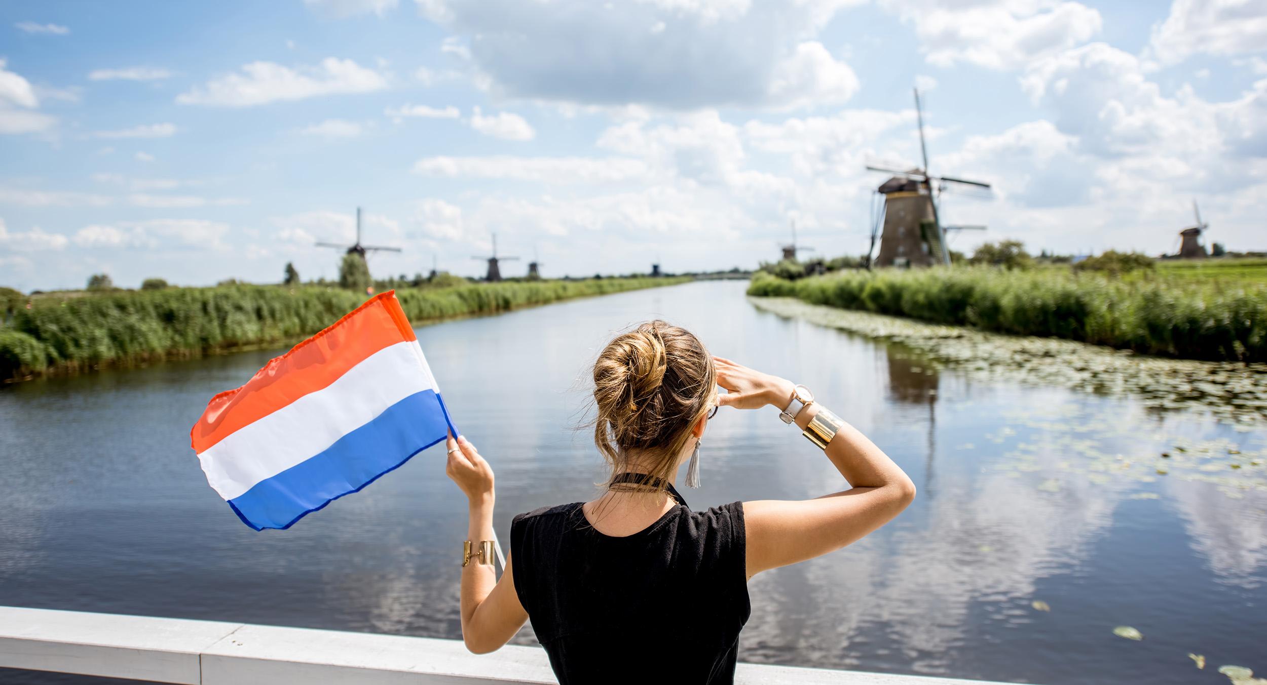 Een vrouw kijkt naar een molen in de verte, met in haar linkerhand een Nederlandse vlag en op de achtergrond een rivier en meer molens