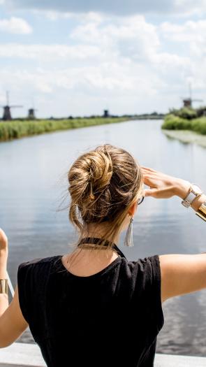 Een vrouw kijkt naar een molen in de verte, met in haar linkerhand een Nederlandse vlag en op de achtergrond een rivier en meer molens