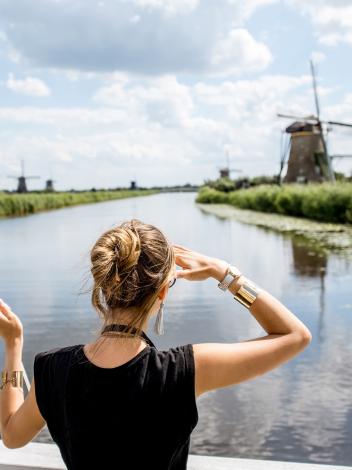 Een vrouw kijkt naar een molen in de verte, met in haar linkerhand een Nederlandse vlag en op de achtergrond een rivier en meer molens