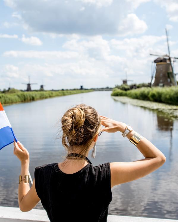 Een vrouw kijkt naar een molen in de verte, met in haar linkerhand een Nederlandse vlag en op de achtergrond een rivier en meer molens