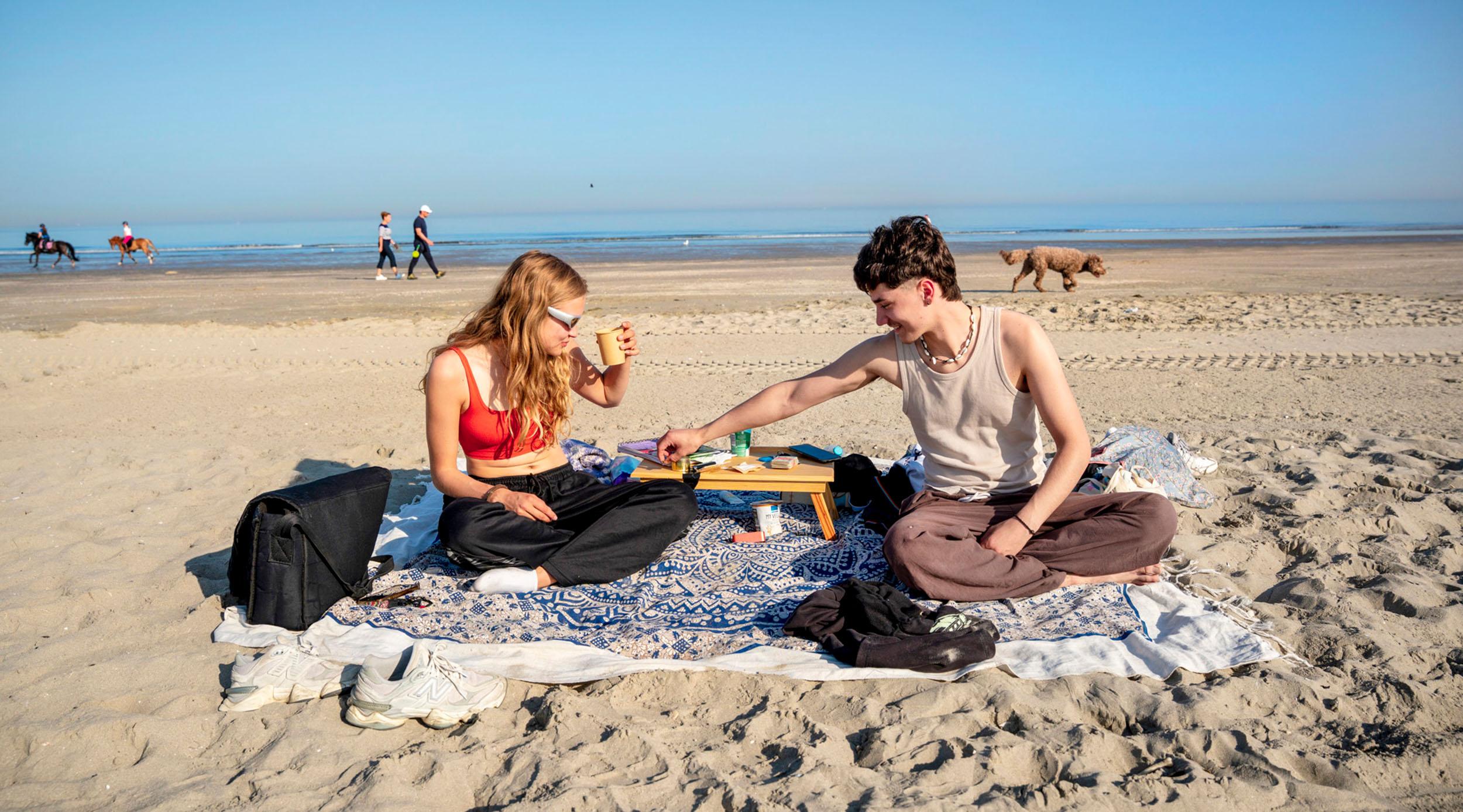Twee jongeren op kleedjes op het strand, op een zonnige dag met de zee op de achtergrond