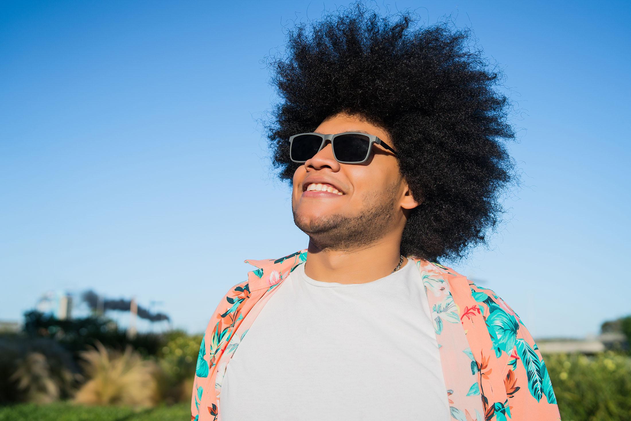 Een man staat lachend naar de lucht te kijken met zijn zonnebril op en zomerse kleding aan