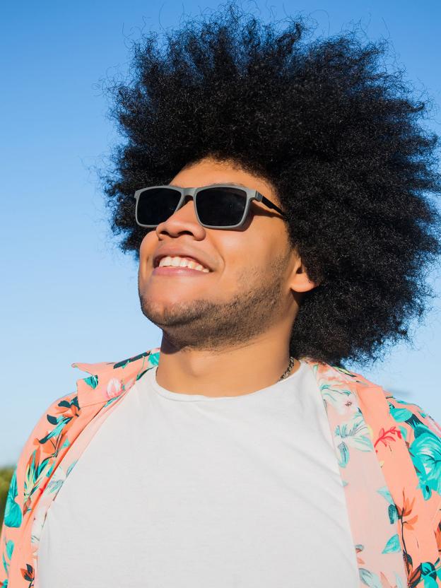 Een man staat lachend naar de lucht te kijken met zijn zonnebril op en zomerse kleding aan