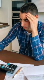 Een man met zijn hoofd in zijn hand, kijkend naar papieren en een rekenmachine aan de keukentafel