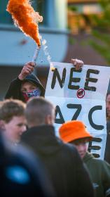 Jongen staat met bord met tekst 'Nee AZC Nee' en oranje rookfakkel in zijn hand met capuchon op