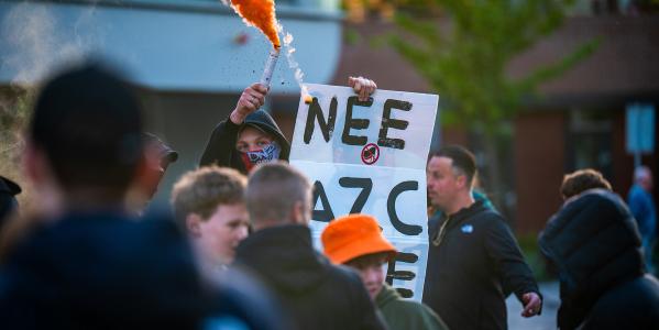 Jongen staat met bord met tekst 'Nee AZC Nee' en oranje rookfakkel in zijn hand met capuchon op