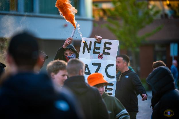 Jongen staat met bord met tekst 'Nee AZC Nee' en oranje rookfakkel in zijn hand met capuchon op