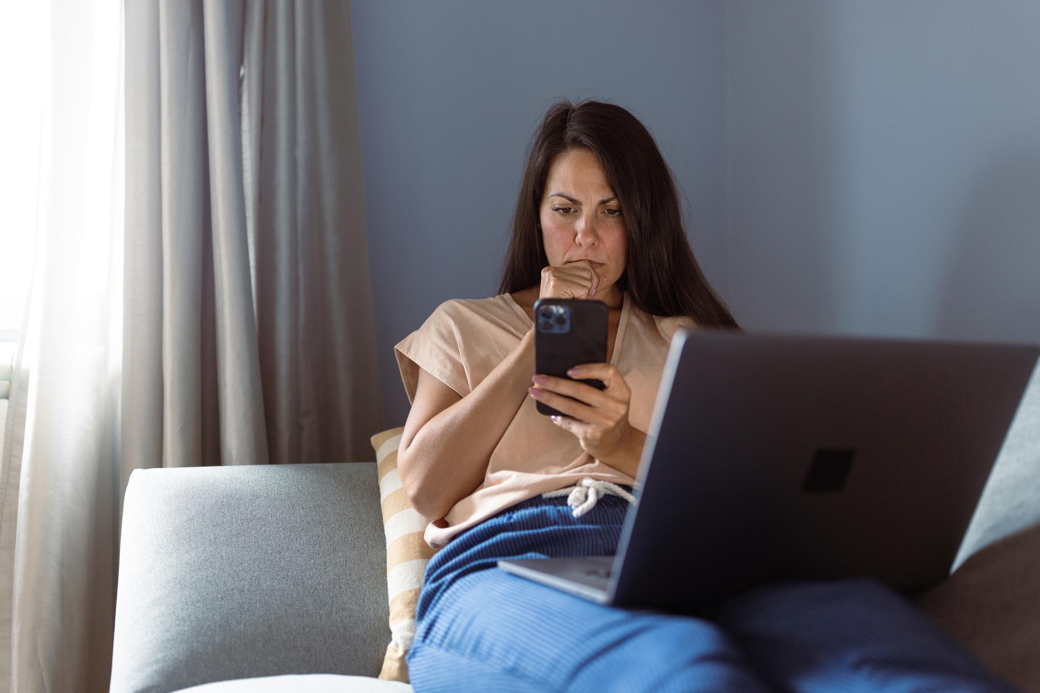 Iemand ligt op de bank met haar laptop op schoot en kijkt met een serieuze blik naar haar telefoon