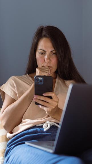 Iemand ligt op de bank met haar laptop op schoot en kijkt met een serieuze blik naar haar telefoon 