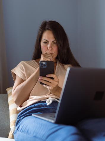 Iemand ligt op de bank met haar laptop op schoot en kijkt met een serieuze blik naar haar telefoon 