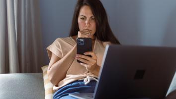 Iemand ligt op de bank met haar laptop op schoot en kijkt met een serieuze blik naar haar telefoon 