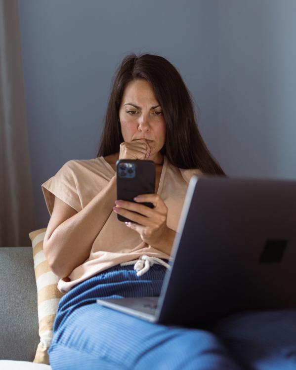 Iemand ligt op de bank met haar laptop op schoot en kijkt met een serieuze blik naar haar telefoon