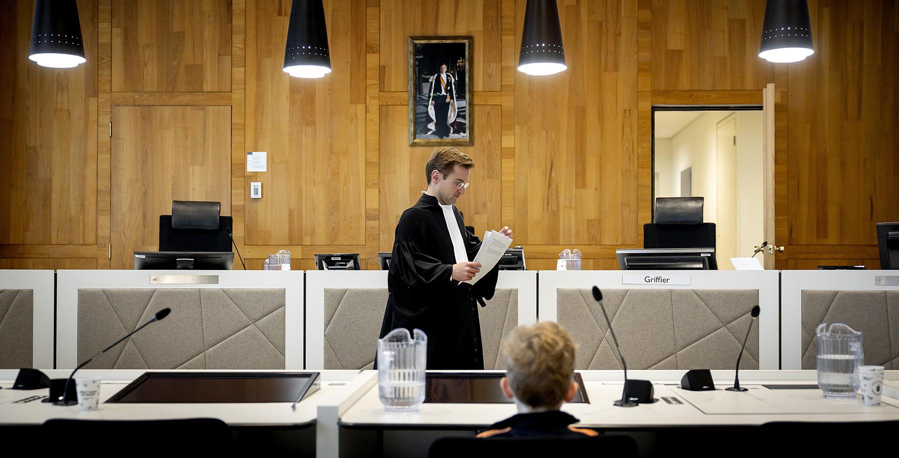 Een man in toga loopt door een rechtszaal met een stapel papieren in zijn hand