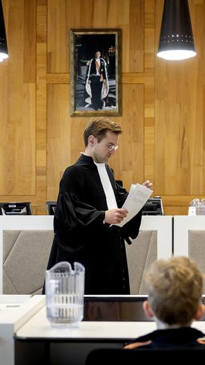 Een man in toga loopt door een rechtszaal met een stapel papieren in zijn hand