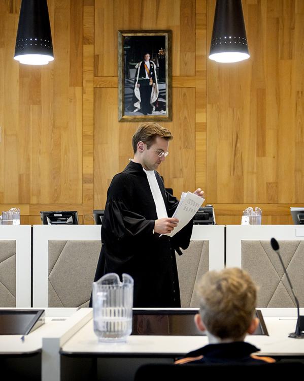 Een man in toga loopt door een rechtszaal met een stapel papieren in zijn hand