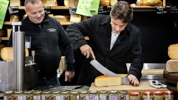 Twee mannen staan achter de toonbank in een kaaswinkel en snijden een punt van een kaaswiel