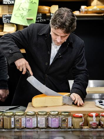 Twee mannen staan achter de toonbank in een kaaswinkel en snijden een punt van een kaaswiel