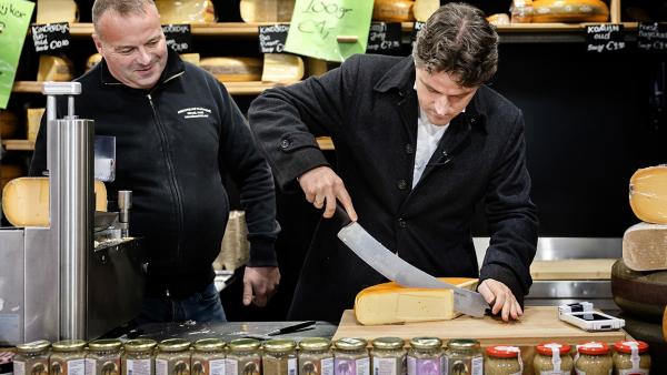 Twee mannen staan achter de toonbank in een kaaswinkel en snijden een punt van een kaaswiel