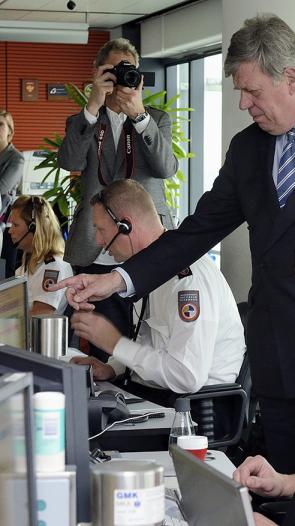 Mensen met koptelefoon op zitten achter computerschermen in de meldkamer. Een man in pak kijkt met een van de schermen mee en wijst ernaar.  