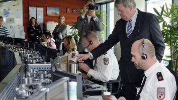 Mensen met koptelefoon op zitten achter computerschermen in de meldkamer. Een man in pak kijkt met een van de schermen mee en wijst ernaar.  