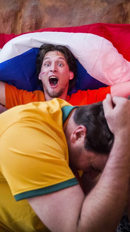Jongen met Nederlandse vlag juicht uitbundig terwijl jongen in Braziliaans voetbalshirt voor hem in elkaar duikt.