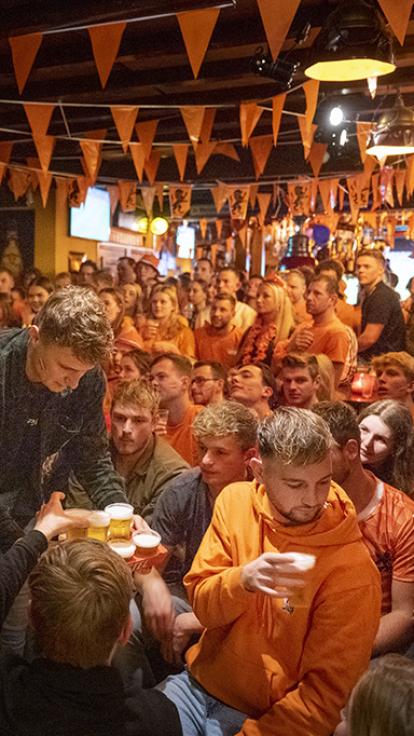 Bomvolle kroeg vol mensen in oranje kleding die gespannen dezelfde kant op kijken. Er loopt ook een jongen rond met een dienblad bier en er hangen oranje vlaggetjes aan het plafond.