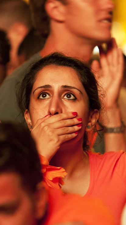 Vrouw in oranje shirt kijkt geschokt met hand voor haar mond omhoog