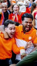 Plein vol juichende blije mensen in oranje kleding. Vooraan staan twee mannen blij te schreeuwen met arm om elkaar heen