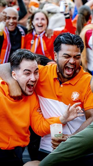 Plein vol juichende blije mensen in oranje kleding. Vooraan staan twee mannen blij te schreeuwen met arm om elkaar heen