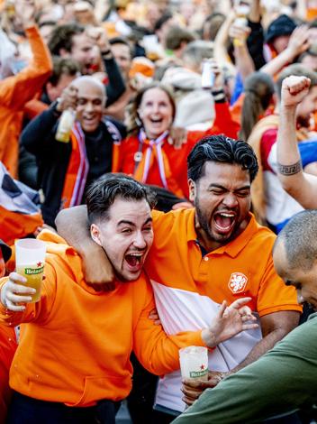 Plein vol juichende blije mensen in oranje kleding. Vooraan staan twee mannen blij te schreeuwen met arm om elkaar heen
