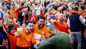 Plein vol juichende blije mensen in oranje kleding. Vooraan staan twee mannen blij te schreeuwen met arm om elkaar heen