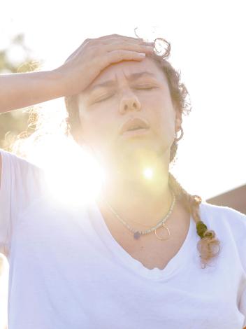 Een vrouw heeft last van de hitte en legt een hand op haar hoofd, met een felle zon achter haar