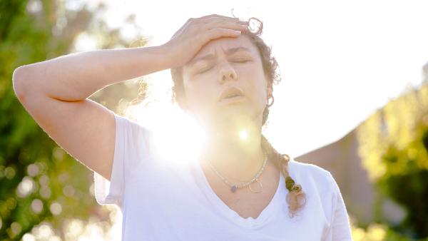Een vrouw heeft last van de hitte en legt een hand op haar hoofd, met een felle zon achter haar