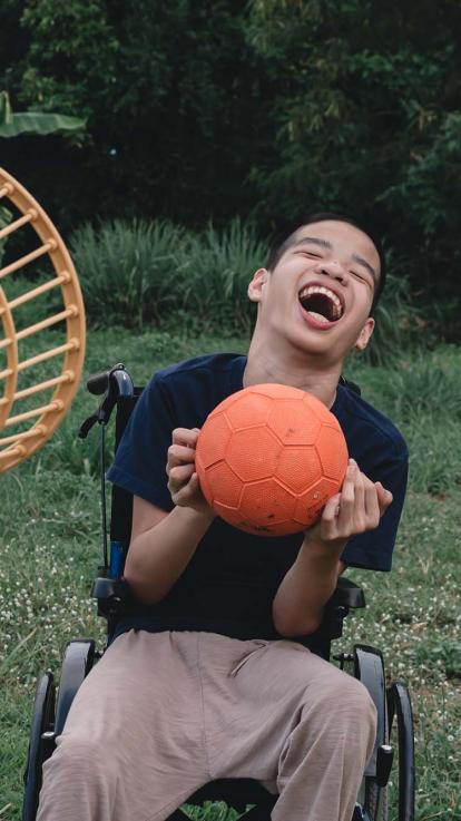 Een jongen in een rolstoel breed lachend met een oranje bal in zijn handen