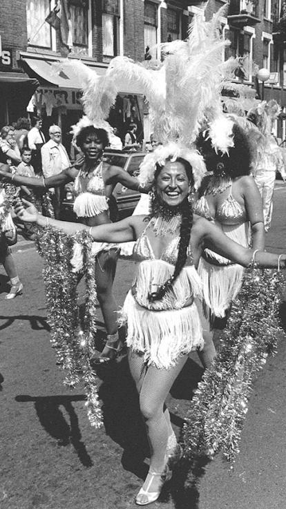 Zwart wit foto van zomercarnaval 1984 waar blije vrouwen in kleding met franjes en veren op hun hoofd dansen op straat