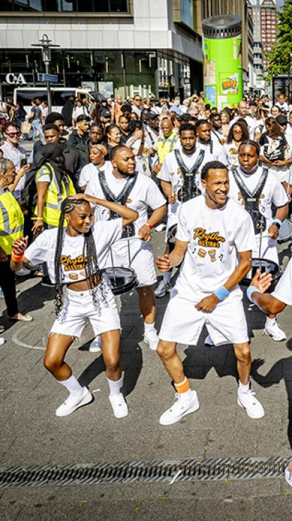 Groep drummers en dansers in witte kleding danst en maakt muziek op straat met veel publiek langs de zijlijn. Voorop loopt een man te begeleiden achteruit met rug richting de camera.