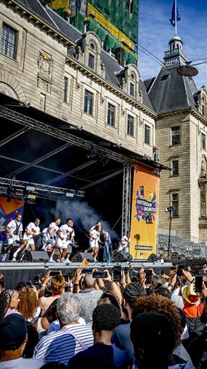 Plein in Rotterdam met podium waar brassband staat te drummen. Veel mensen uit het publiek staat te filmen. 