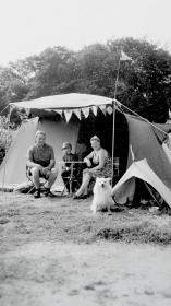 Een zwart-witfoto van een gezin met hond die voor hun tent zitten