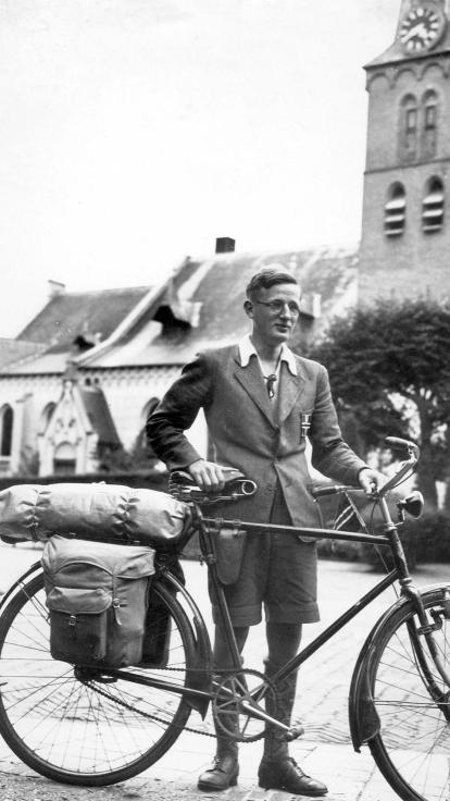 Een zwart-witfoto van een man die poseert met zijn fiets, tent achterop, op een pleintje 