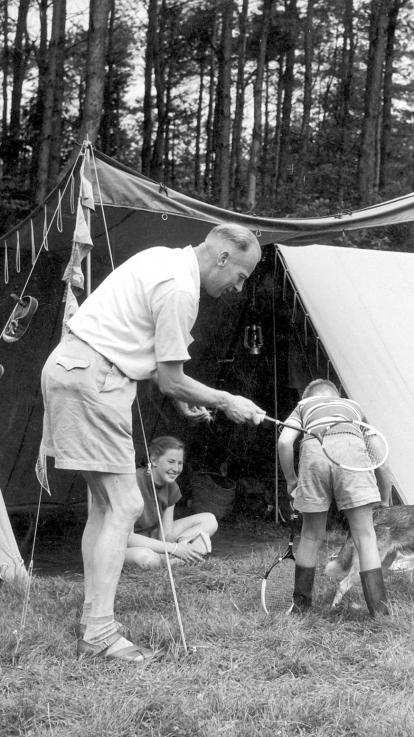Zwart-witfoto van familie aan het kamperen. De vader houdt zijn tennisracket achter de kont van zijn bukkende zoon.