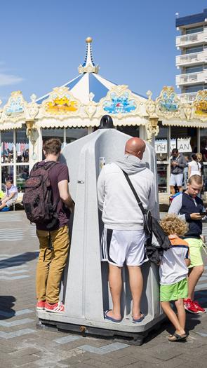 Op de scheveningse boulevard staan urinoirs waar 3 mannen aan het plassen zijn. Daarnaast lopen kindjes. 